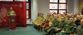 Na zdjęciu widać oficjalne spotkanie przedstawicieli służb mundurowych. Na pierwszym planie siedzą funkcjonariusze w mundurach wojskowych i strażackich, a w tle osoba w mundurze Państwowej Straży Pożarnej przemawia przy mównicy. Na czerwonym tle za nim widnieje napis "Państwowa Straż Pożarna" oraz logo PSP. Sala jest dobrze oświetlona, z dużymi oknami i zieloną wykładziną.