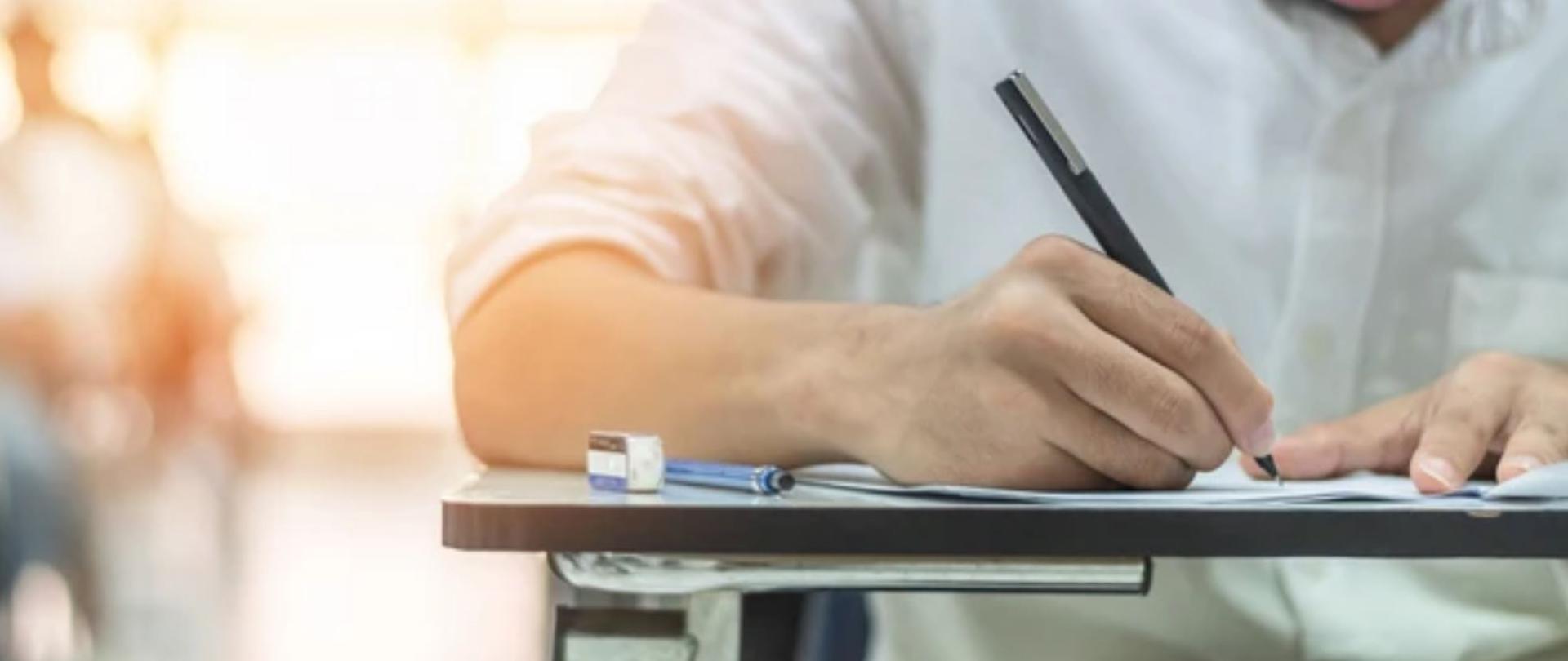 Uczeń w białęj koszuli pisze egzamin przy stoliku, trzyma w reku długopis. 