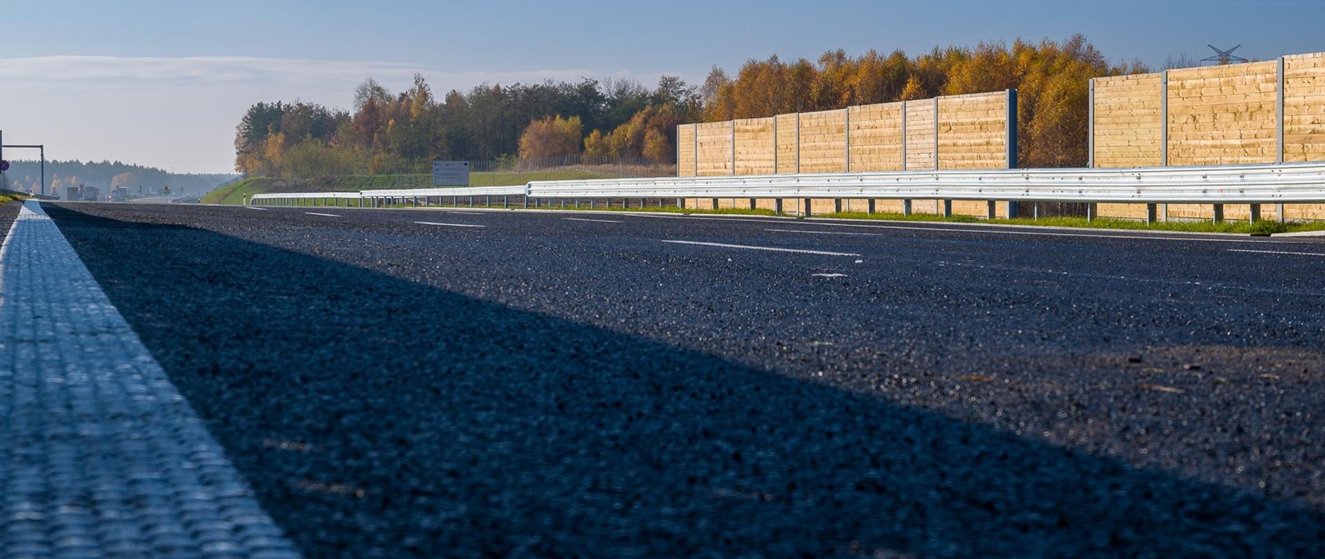 Słoneczny dzień. Zbliżenie na jezdnię autostrady A1. W tle widoczne bariery, a za nimi drzewa.