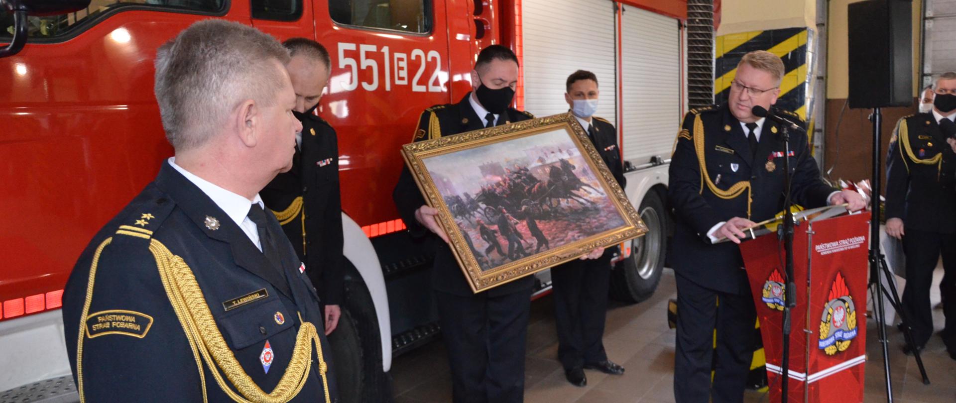 Zdjęcie przedstawia komendanta wojewódzkiego przekazującego obraz dla odchodzącego komendanta powiatowego, widoczny także nowo powołany komendant powiatowy oraz naczelnik wydziału kadrowego w komendzie wojewódzkiej psp w łodzi, wszyscy ubrani w mundury wyjściowe.