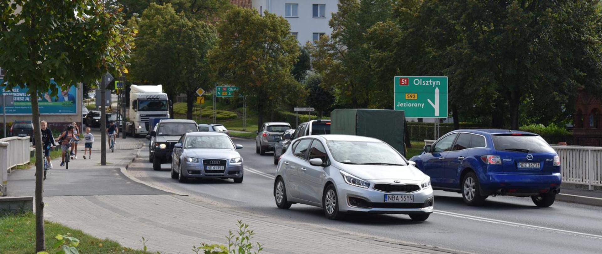 Na zdjęciu widoczna jest ruchliwa ulica, po której porusza się kilka pojazdów, w tym biały samochód na pierwszym planie, niebieski samochód oraz ciężarówka. Po prawej stronie znajduje się zielony znak drogowy wskazujący kierunki do Olsztyna i Jezioran drogą krajową nr 51. W tle widać drzewa, zabudowania oraz wieżę z zegarem. Po lewej stronie ulicy spacerują piesi i poruszają się rowerzyści.