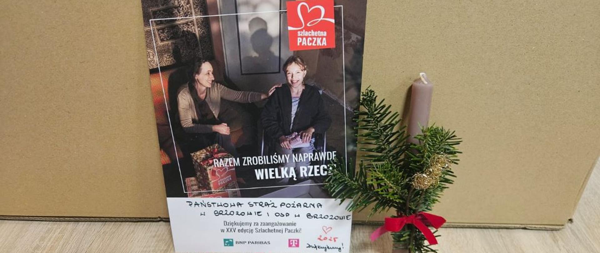 Kolorowa fotografia przedstawiająca promocję akcji „Szlachetna Paczka” z podpisem ręcznym u dołu „Państwowa Straż Pożarna w Brzozowie i OSP Brzozowie”. Z boku znajduje się świeca z igliwiem oraz wstążką.