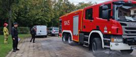 Plac z trelinki na którym stoi ciężkie auto gaśnicze z włączonymi zraszczami, auto obserwuje trzech strażaków, jeden z nich trzyma w ręku notatnik