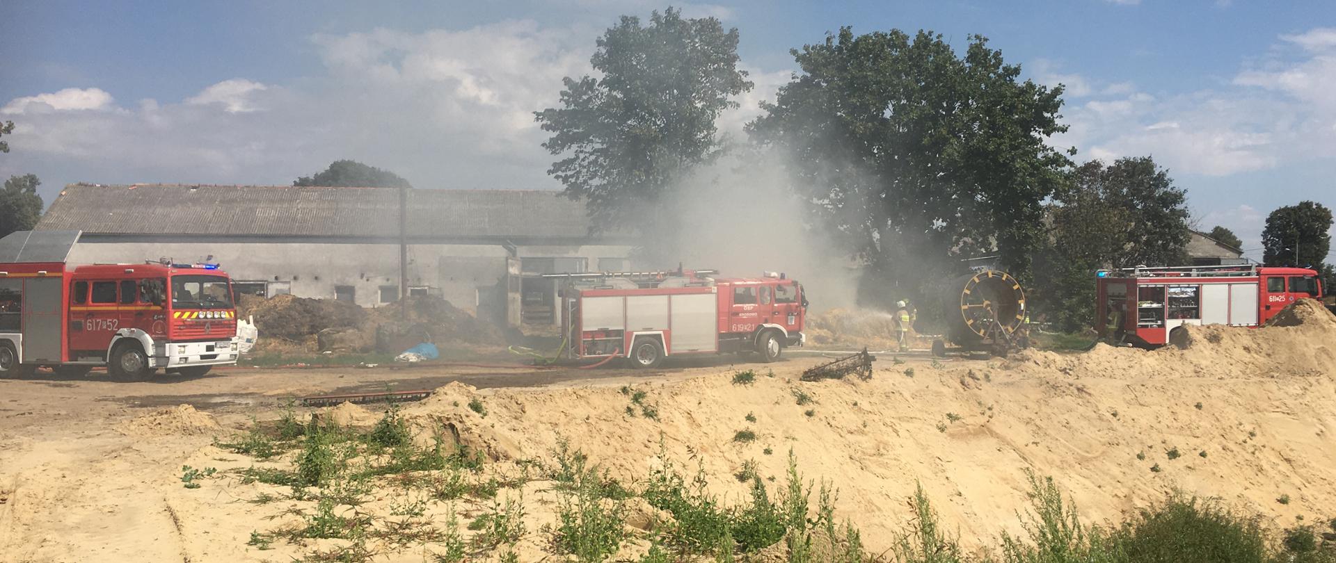 Trzy wozy gaśnicze na tle palących się balotów słomy ułożonych w stóg. Strażacy z linią gaśniczą podają prąd wody na pożar. W powietrzu unosi się dym.
