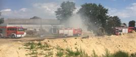 Trzy wozy gaśnicze na tle palących się balotów słomy ułożonych w stóg. Strażacy z linią gaśniczą podają prąd wody na pożar. W powietrzu unosi się dym.