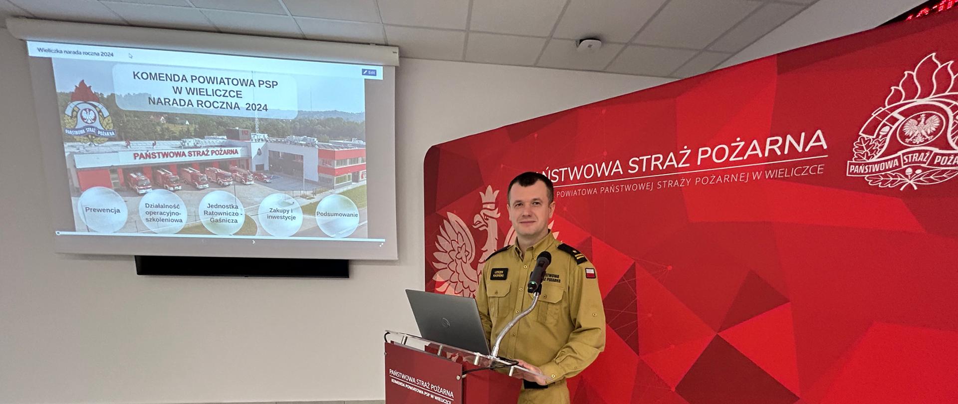 Na zdjęciu komendant powiatowy PSP w Wieliczce mł. bryg. Leszek Kasiński podczas omawiania prezentacji przy mównicy. W tle prezentacja.