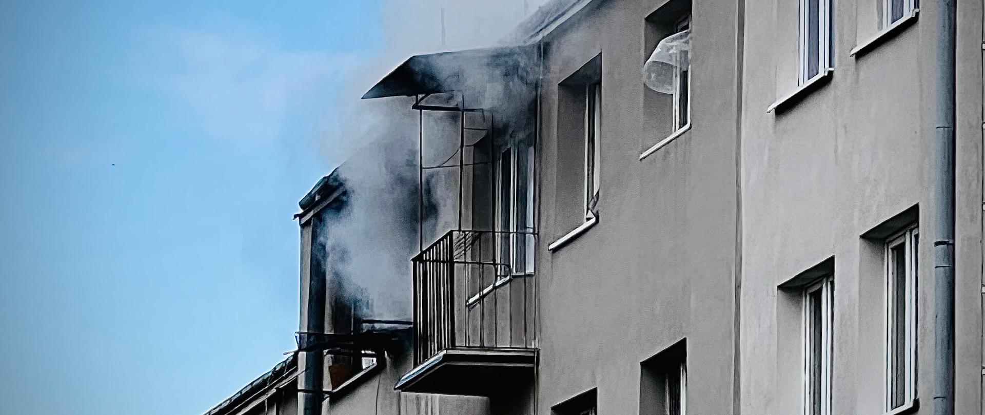 Zdjęcie przedstawia fasadę bloku mieszkalnego. Na ostatniej kondygnacji widoczny jest gęsty dym wydobywający się z okien. W tle widoczne jest błękitne niebo.