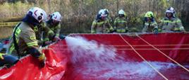 Zdjęcie przedstawia strażaków przy zbiorniku na wodę. Zbiornik jest plastikowy otwarty z góry w kolorze czerwonym. Strażacy nalewają wodę ze zbiornika przeciwpożarowego do zbiornika na brzegu. W oddali widać las. 