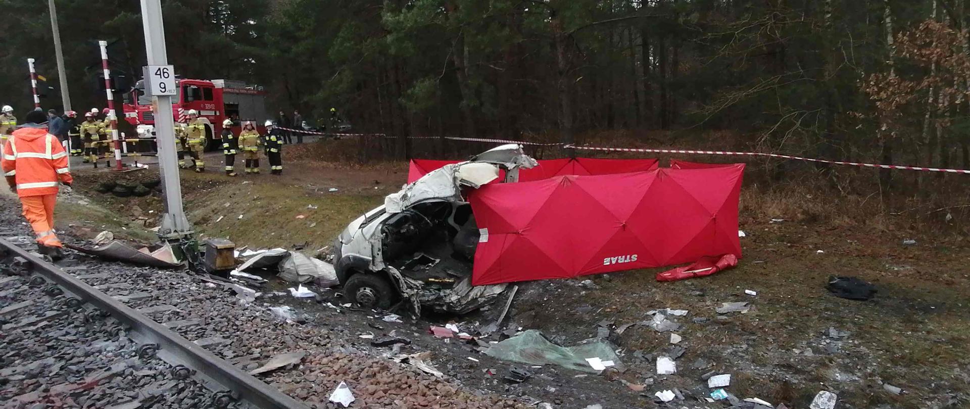 Na zdjęciu przód samochodu po wypadku, teren wokół samochodu częściowo zasłonięty czerwonym parawanem z napisem straż. Po lewej stronie na drodze w lesie stoi wóz strażacki, strażacy oraz osoby cywilne. W lewej dolnej części zdjęcia torowisko przy którym idzie mężczyzna w pomarańczowym ubraniu. 