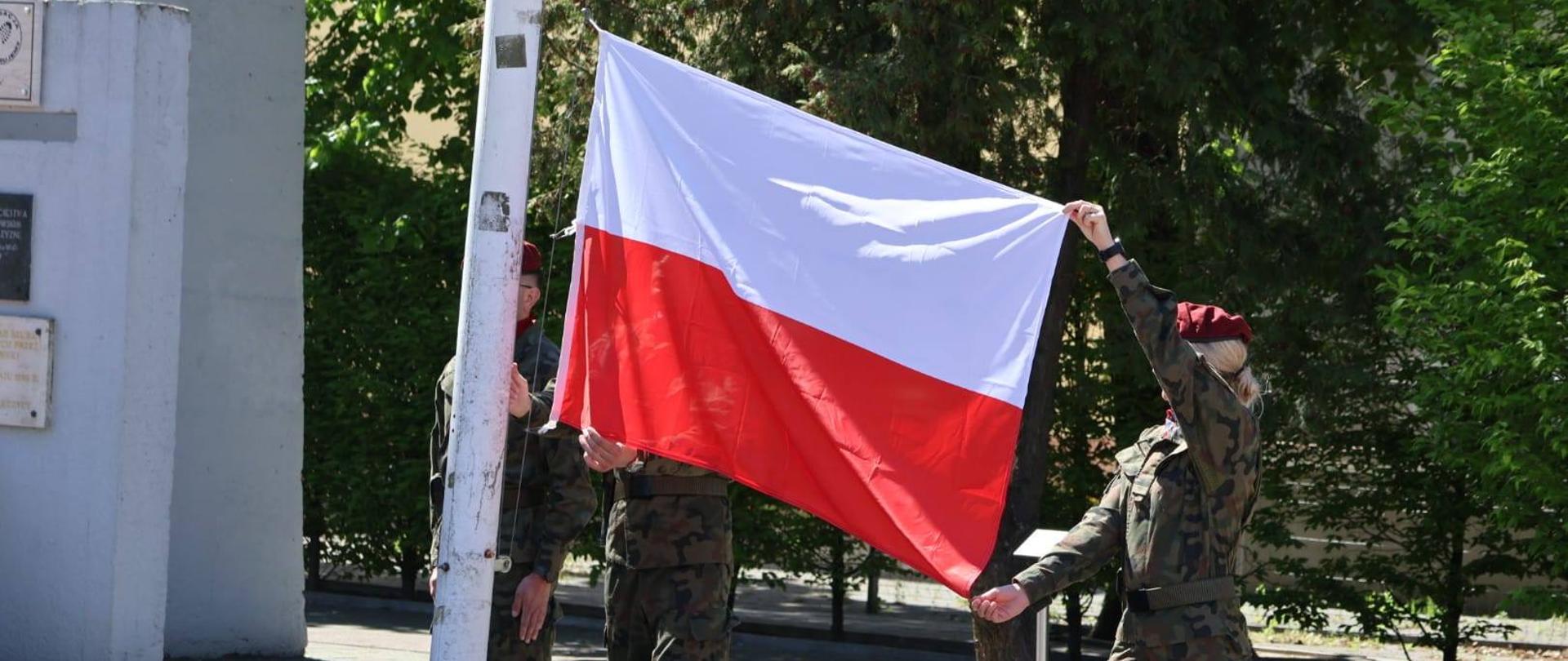 Na zdjęciu widoczna jest ceremonia podnoszenia polskiej flagi na maszt. Trzy osoby w wojskowych mundurach i bordowych beretach uczestniczą w przygotowaniu flagi do podnoszenia.