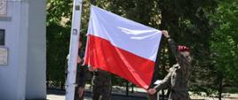 Na zdjęciu widoczna jest ceremonia podnoszenia polskiej flagi na maszt. Trzy osoby w wojskowych mundurach i bordowych beretach uczestniczą w przygotowaniu flagi do podnoszenia.