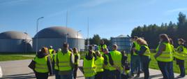 Zdjęcie przedstawia grupę kilkunastu osób uczestniczących w szkoleniu lub wizycie studyjnej na terenie instalacji przemysłowej. Uczestnicy stoją na utwardzonym placu i są zwróceni w kierunku prowadzącego, który znajduje się w centralnej części grupy. Większość osób ma na sobie żółte kamizelki ostrzegawcze z odblaskowymi pasami. W tle widoczne są duże, okrągłe zbiorniki przykryte szarymi kopułami, instalacje technologiczne oraz rurociągi. Teren jest ogrodzony i otoczony zielenią oraz drzewami. Spotkanie odbywa się w słoneczny, bezchmurny dzień.