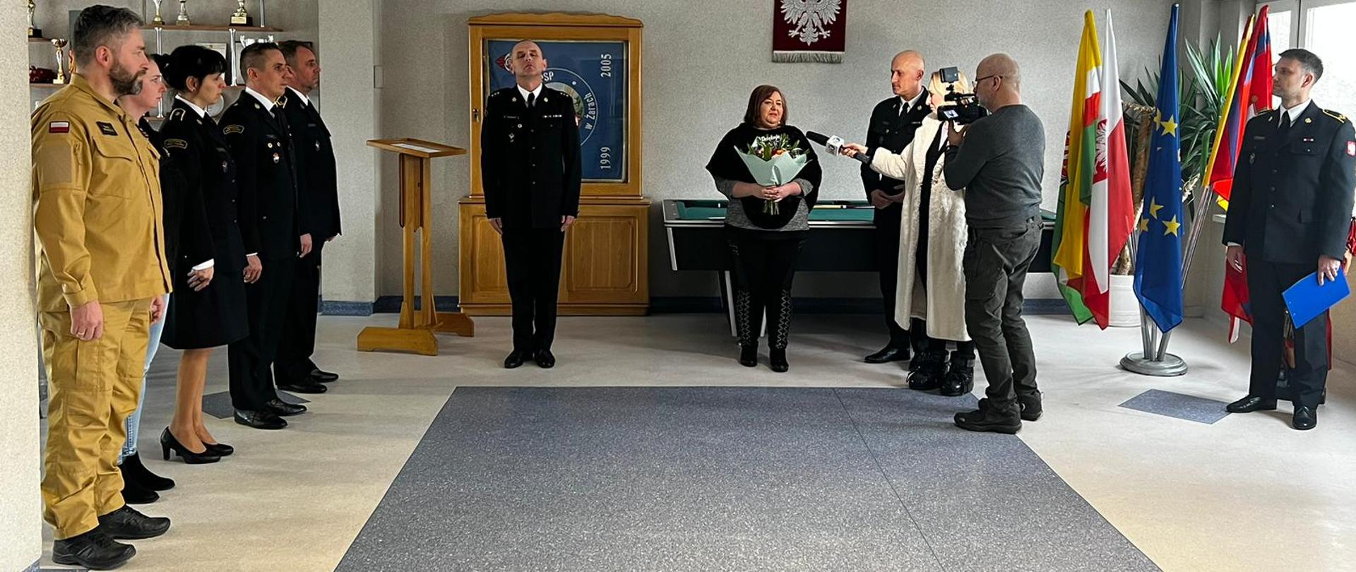 Zdjęcie przedstawia Panią Dorotę, udzielającą wywiadu TV Regionalnej. Obok widoczne są osoby biorące udział w uroczystej zbiórce. 