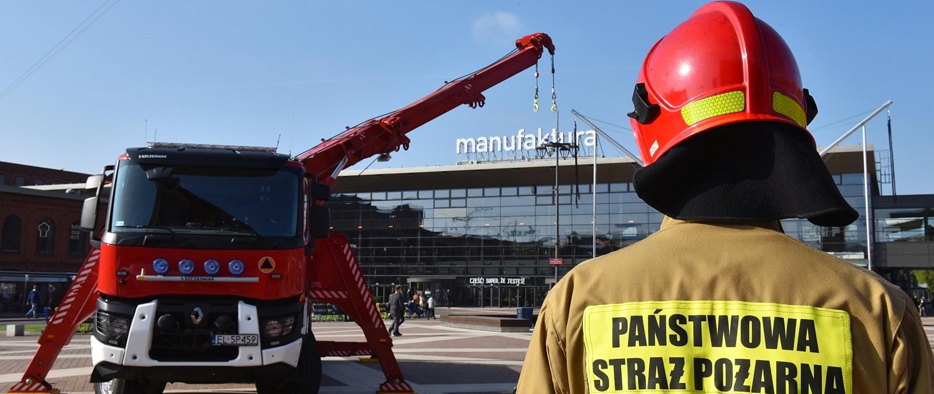 na zdjęciu strażak stojący tyłem widać czerwony hełm oraz ubranie koloru piaskowego z napisem Państwowa straż pożarna, samochód ratownictwa technicznego z żurawiem w tle ściana frontowa centrum handlowego manufaktura w łodzi