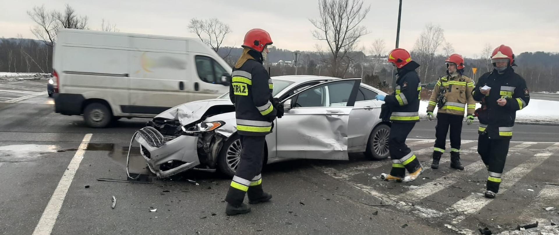 Samochód koloru jasnego biorący udział w zdarzeniu stoi na skrzyżowaniu. Obok niego stoi czterech strażaków. Na dalszym tle samochody uczestniczące w ruchu drogowym..