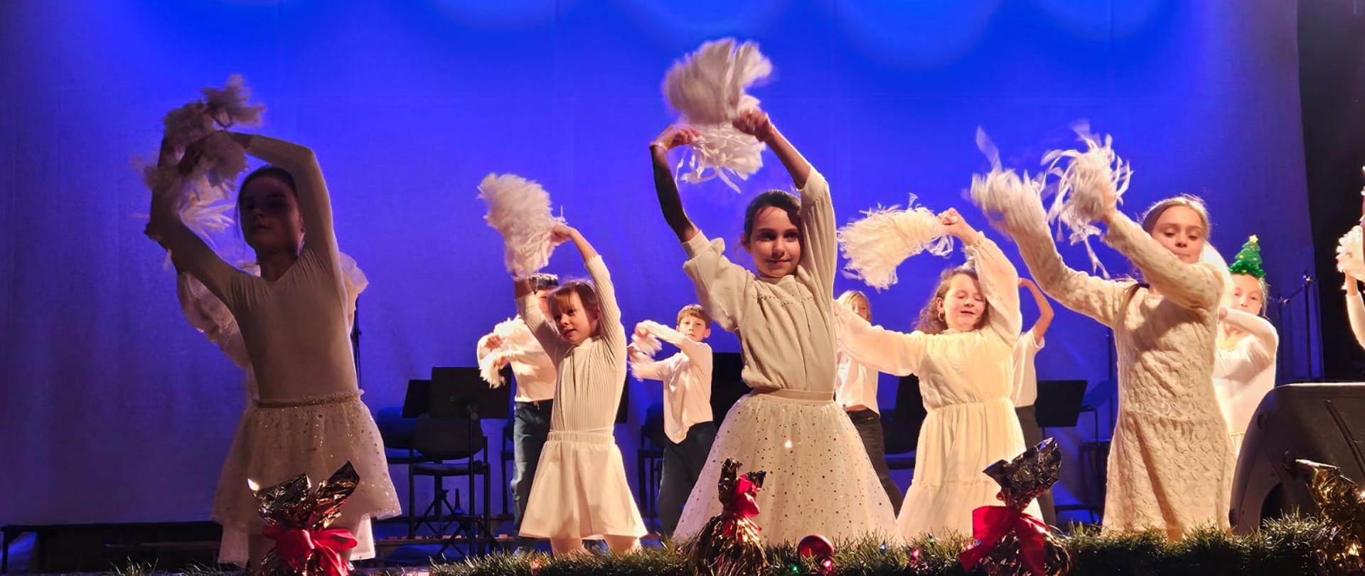 Zdjęcie kolorowe. Grupa dzieci w białych strojach śnieżynek tańczy na scenie na jednolitym niebieskim tle. W rękach trzymają białe, papierowe gwiazdki.