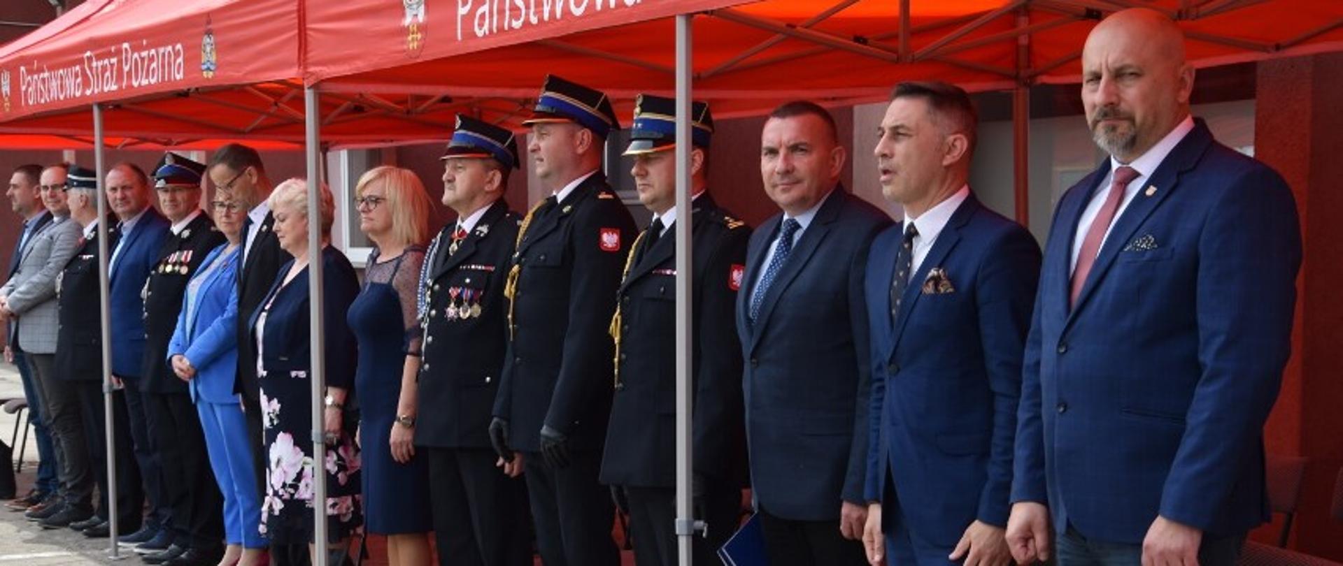 pod czerwonym namiotem stoi grupa mężczyzn w mundurach , garniturach, śród grupy stoją także kobiety 