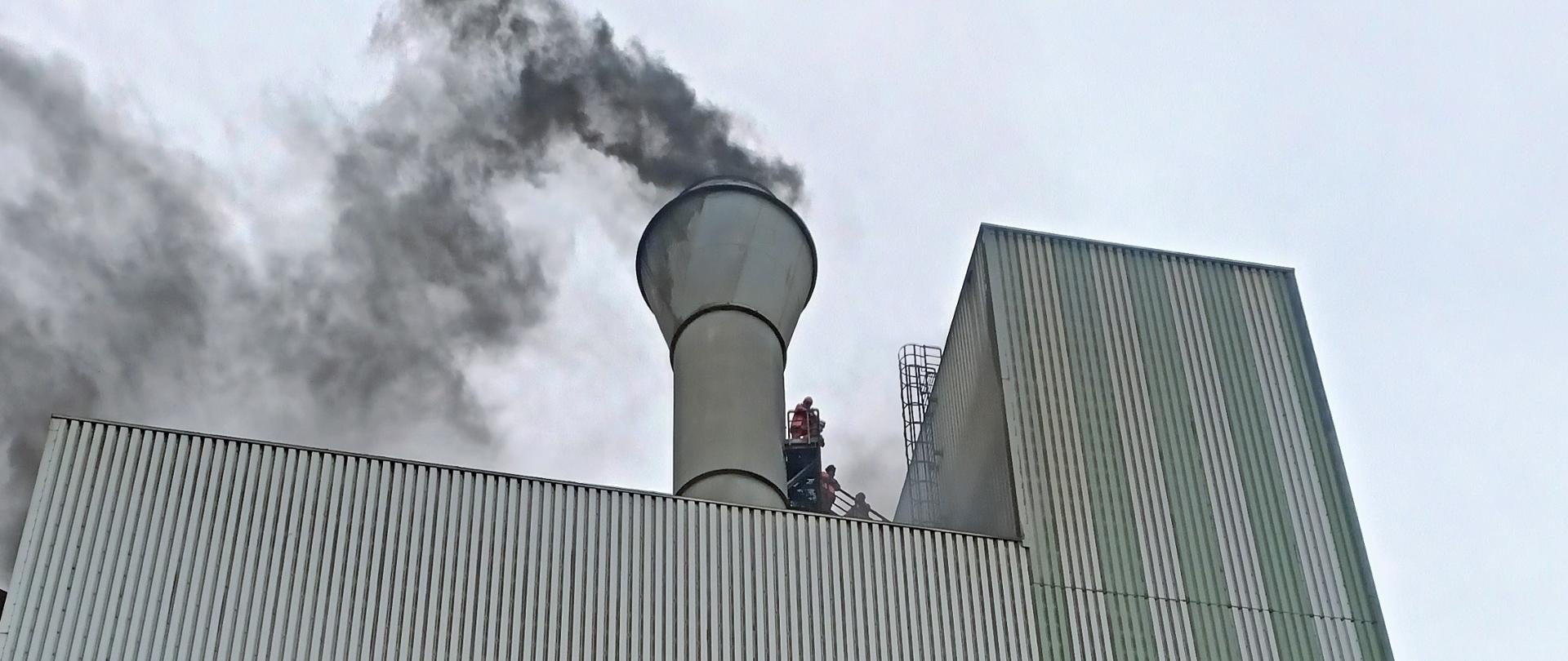 Na zdjęciu widać wysoki budynek przemysłowy koloru szarego o elewacji z blachy trapezowej. Z nad dachem widać unoszący się dym. Na pomoście przy kominie stoją odcięci przez pożar pracownicy. 