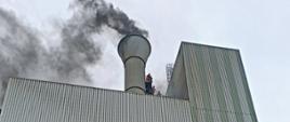 Na zdjęciu widać wysoki budynek przemysłowy koloru szarego o elewacji z blachy trapezowej. Z nad dachem widać unoszący się dym. Na pomoście przy kominie stoją odcięci przez pożar pracownicy. 