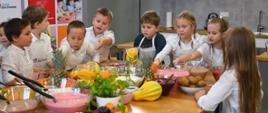 Grupa małych dzieci w białych fartuszkach stoi dookoła stołu, na którym jest dużo kolorowych owoców.