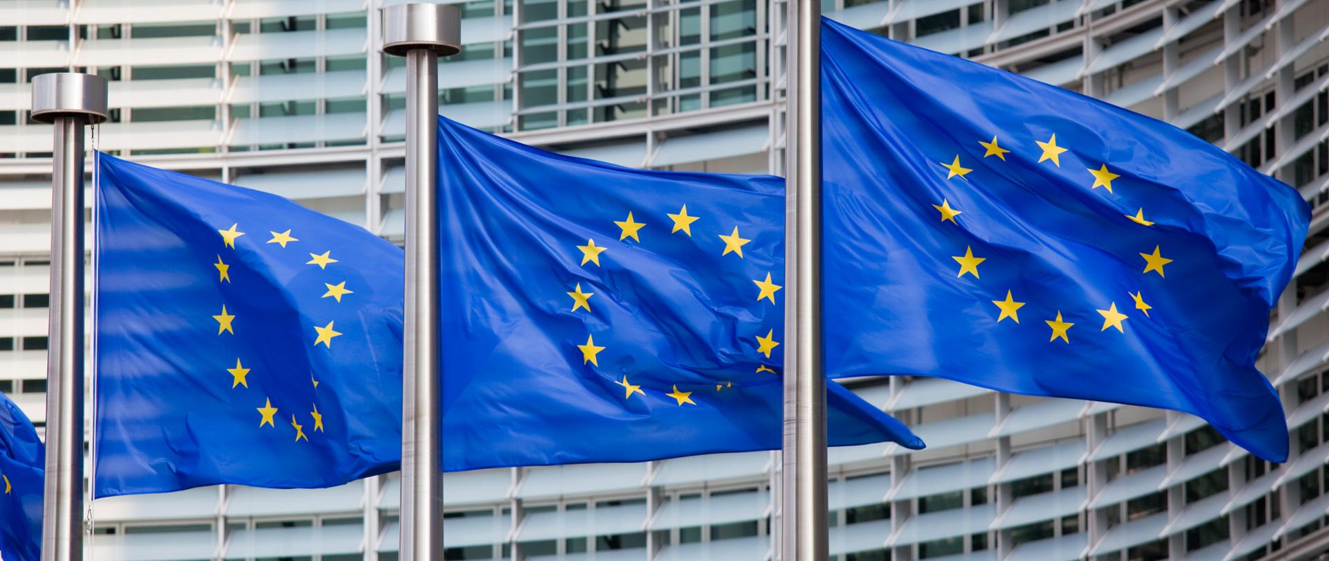 Zdjęcie w poziomie. Trzy flagi Unii Europejskiej w rzędzie na tle parlamentu Europejskiego