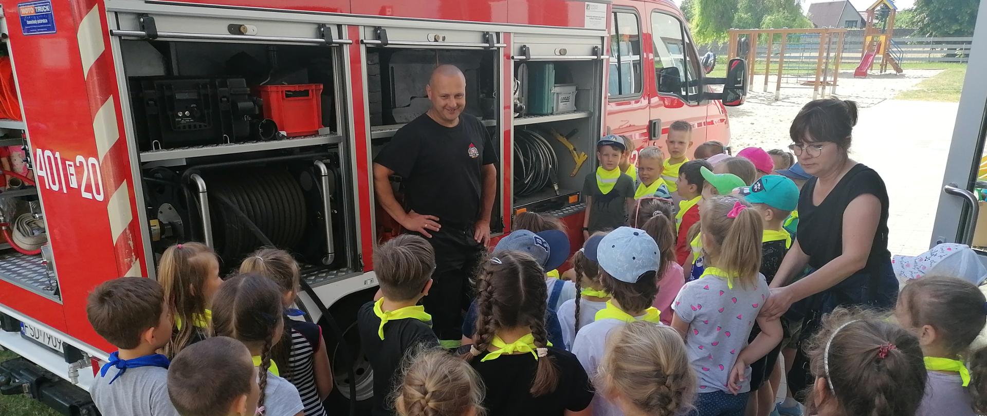 Fotografia przedstawia ujęcie samochodu pożarniczego stojącego prawym bokiem i częściowo tyłem do fotografującego. Skrytki pojazdu są pootwierane, przed pojazdem stoi strażak w ubraniu koszarowym. Przed strażakiem stoi półkole dzieci w wieku przedszkolnym wraz z opiekunką. Dzieci stoją tyłem do fotografującego. Jest dzień, słonecznie, w tle widoczny plac zabaw.