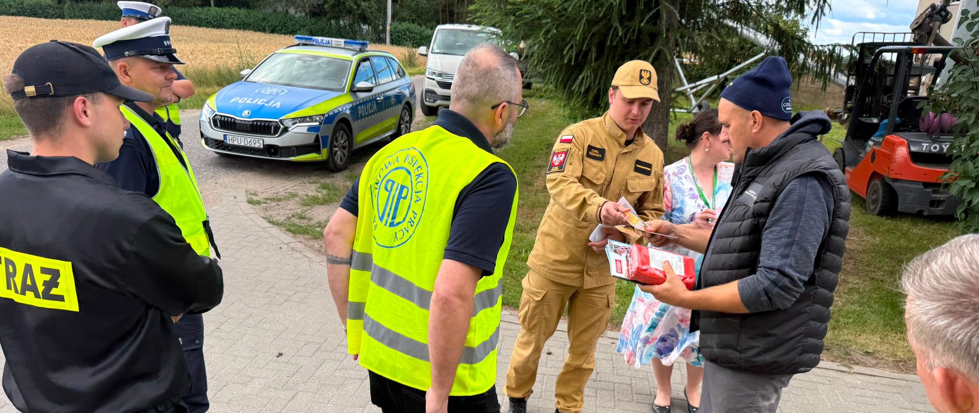 Na zdjęciu służby biorące udział w akcji „Bezpieczne Żniwa” rozmawiają z rolnikiem na utwardzonym podjeździe przed posesją. Strażak przekazuje mężczyźnie ulotkę z informacjami dotyczącymi usytuowania stert i stogów, a obok stoją policjanci oraz przedstawiciel inspekcji pracy. W tle widoczny jest radiowóz policyjny, pojazdy oraz drzewa i pola.