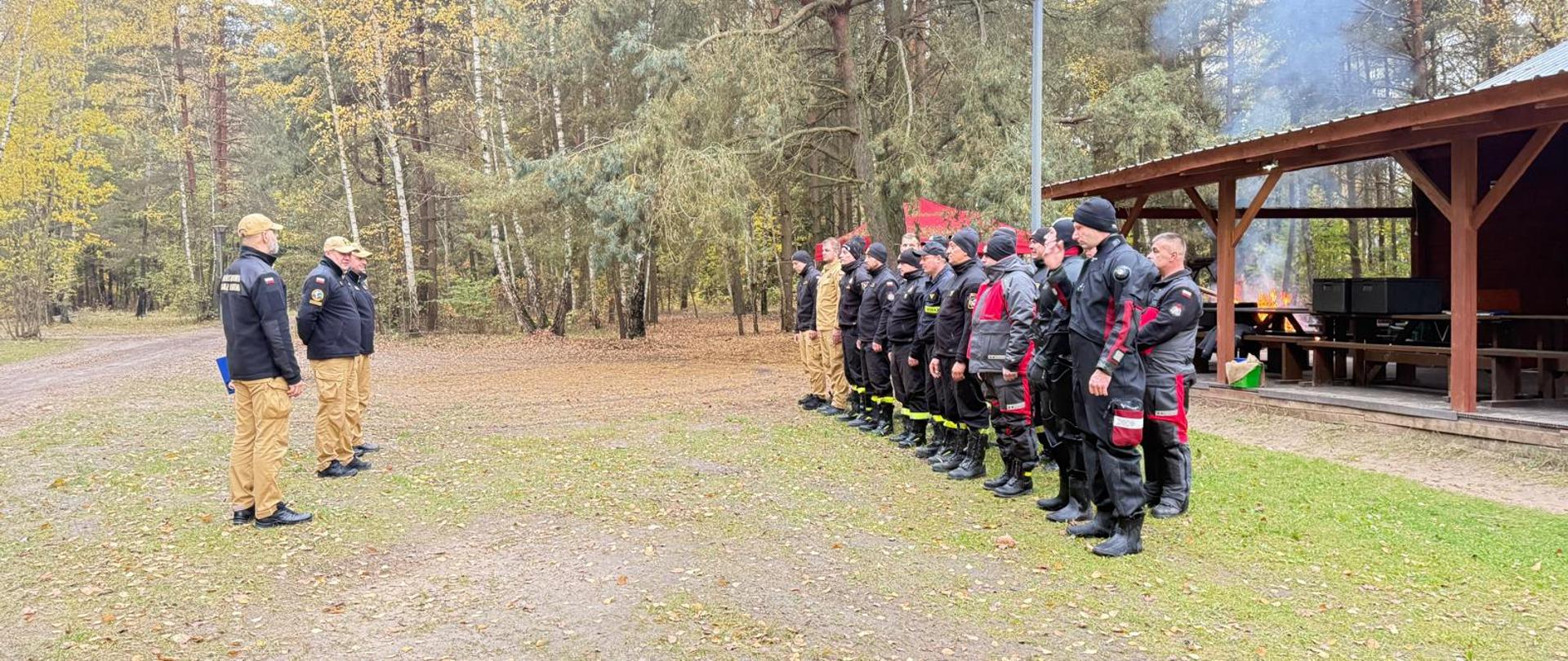 Strażacy stoją w dwuszeregu. Przed nimi, w odległości ok. 5 metrów stoi trzech strażaków. Wszyscy stoją na polanie. Za strażakami stojącymi w dwuszeregu, po prawej stronie zdjęcia widać fragment drewnianego budynku. W tle i ciemnozielone i lekko pożółkłych drzew. 