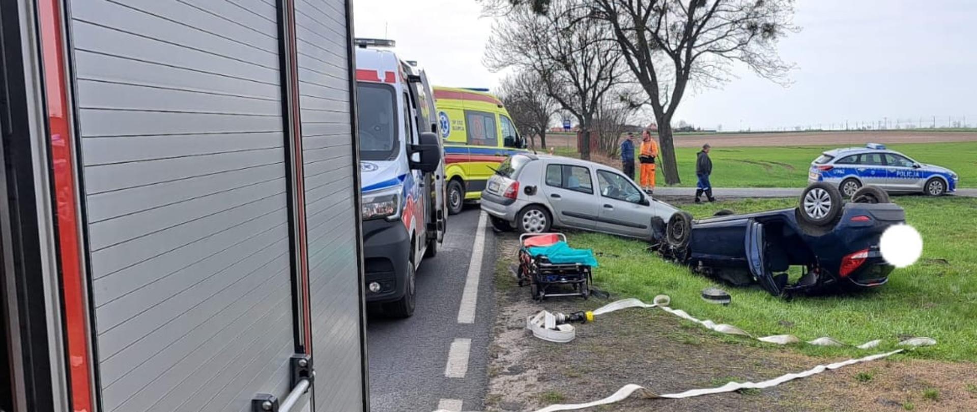 Zdjęcie przedstawia od lewej strony: bok pojazdu straży pożarnej, i dwóch karetek. Po prawej stronie od nich znajdują się dwa samochody osobowe po wypadku- jeden na dachu, drugi na kołach. 