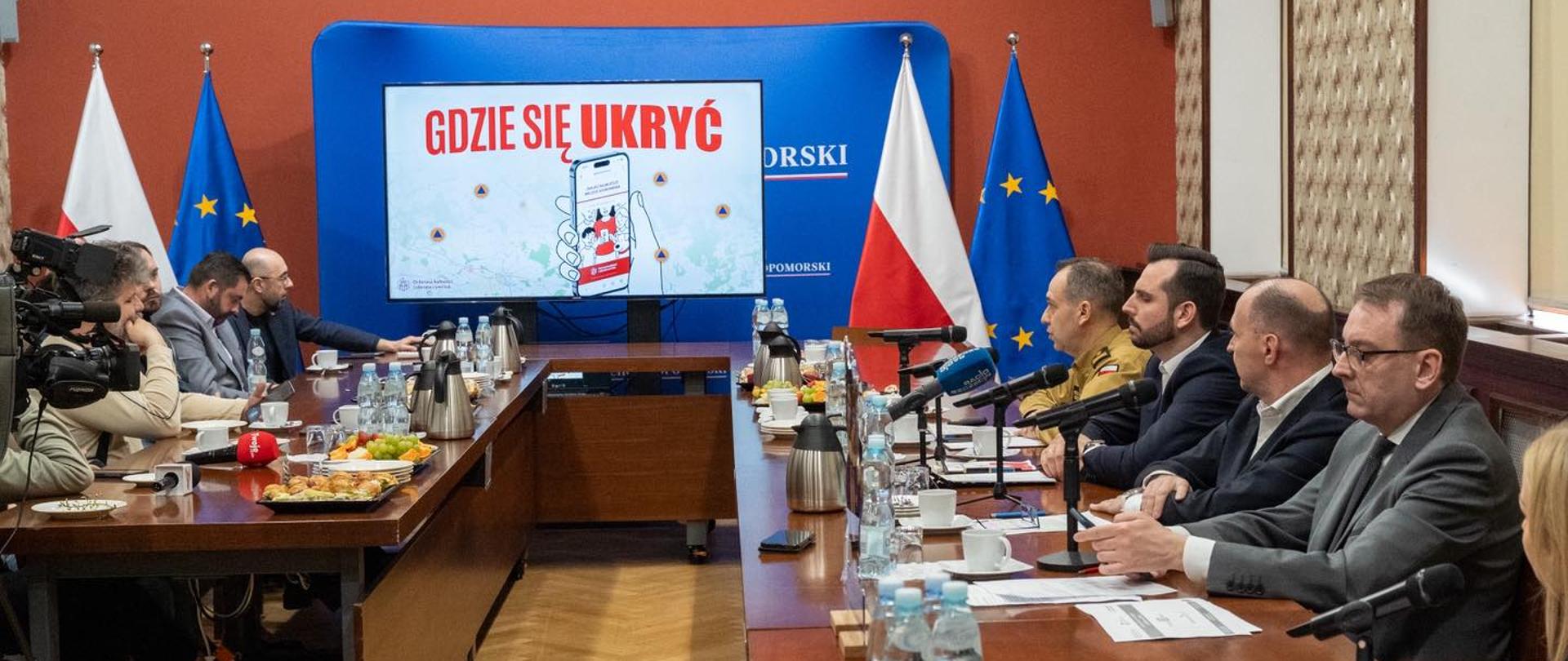 W sali konferencyjnej odbywa się spotkanie z udziałem przedstawicieli instytucji publicznych i mediów. Uczestnicy siedzą po obu stronach długiego stołu z mikrofonami i dokumentami. Na ekranie wyświetlana jest prezentacja z napisem w języku polskim. W tle widoczne są flagi Polski i Unii Europejskiej.