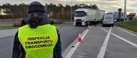 Na pierwszym planie: umundurowany funkcjonariusz lubuskiej Inspekcji Transportu Drogowego stoi i trzyma w prawej ręce alkomat przesiewowy. W tle: trzy ciężarówki zatrzymane do kontroli trzeźwości wjeżdżają do punktu kontrolnego przy trasie.