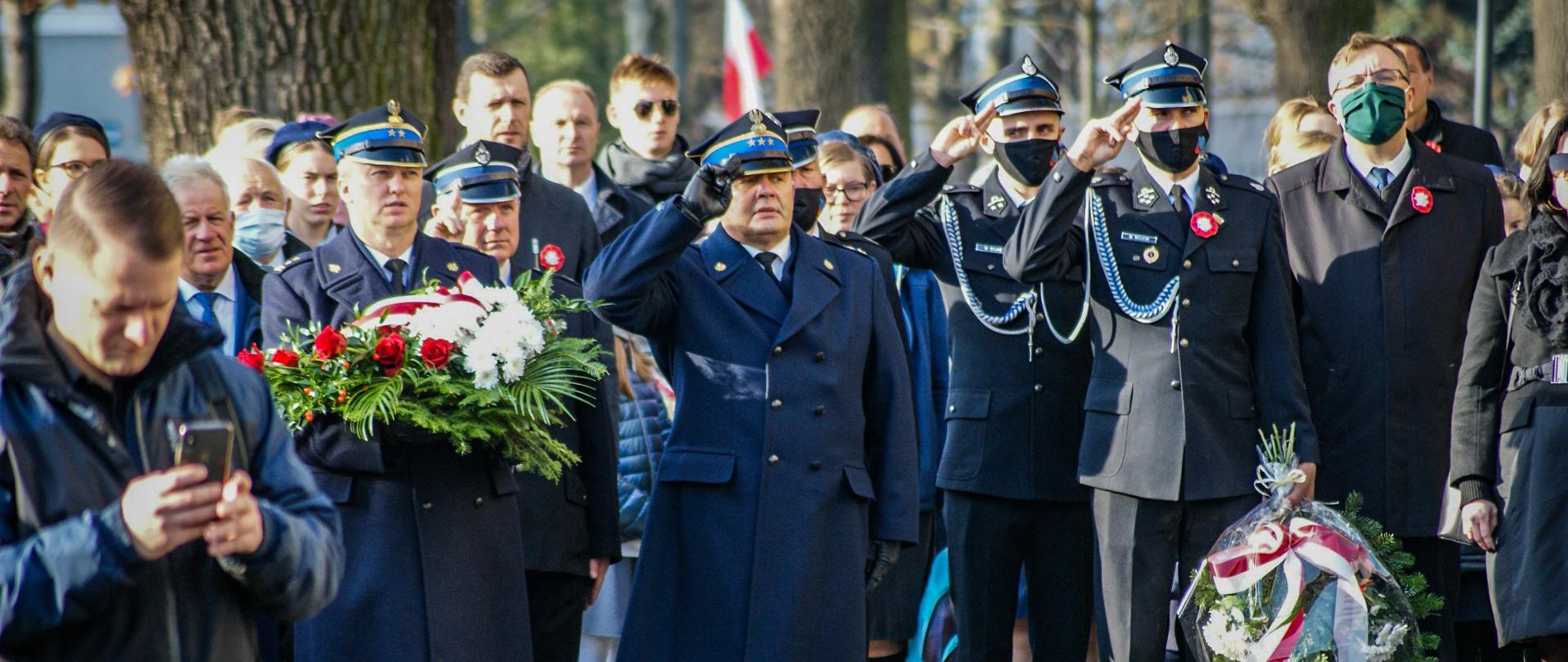Fotografia przedstawia uczestników obchodów podczas śpiewania hymnu państwowego, umundurowani funkcjonariusze oddają honory poprzez salutowanie. Na pierwszym planie osoba nagrywająca uroczystość smartfonem. Dalej strażacy w mundurach wyjściowych lub płaszczach koloru granatowego.