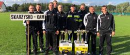 Zdjęcie obrazuje strażaków ze Skarżyska-Kamiennej pozujących do grupowego zdjęcia wraz z Komendantem Powiatowym, po ceremonii wręczenia nagród i pucharów. Przed nimi stoją nagrody jakie wygrali strażacy w konkurencjach indywidualnych oraz pamiątkowe puchary, których jest ich aż osiem. Strażacy stoją na płycie boiska do piłki nożnej, w tle widać drzewa i budynek.