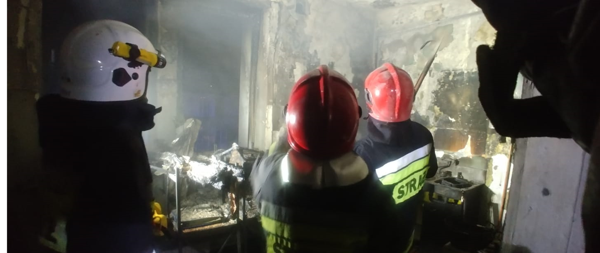 Pomieszczenie zniszczone w wyniku pożaru. W pomieszczeniu trzech strażaków w umundurowaniu wyjściowym.