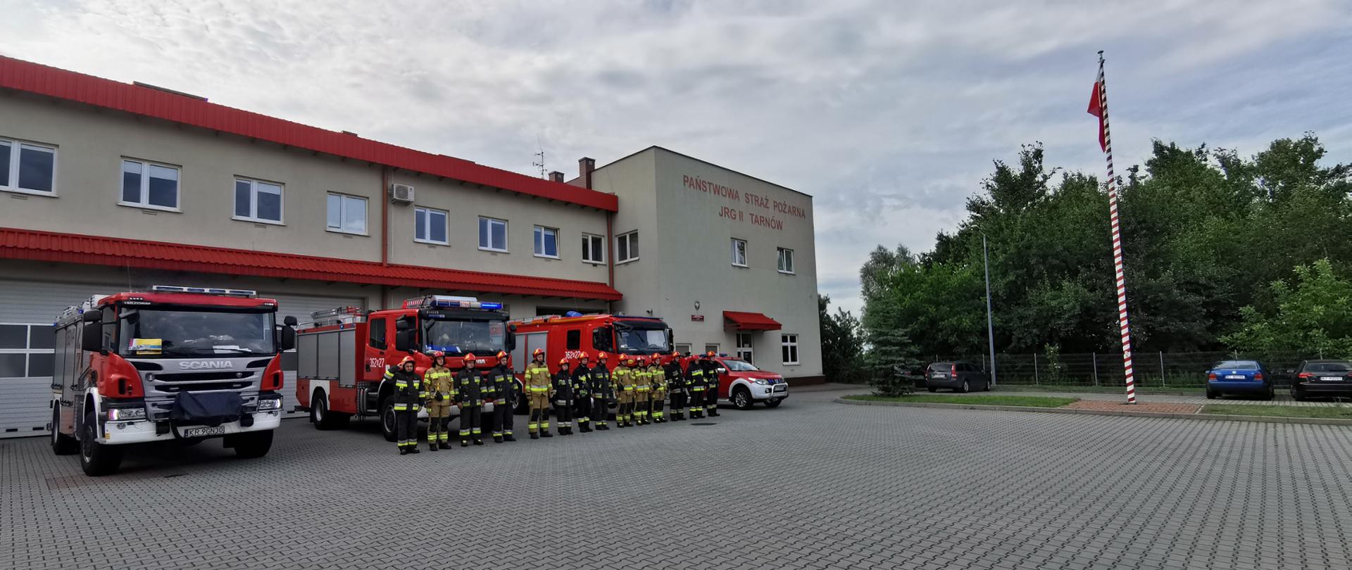 Na zdjęciu widać strażaków na tle 4 samochodów ratowniczo gaśniczych z włączonymi sygnałami ratowniczo gaśniczymi. Samochody ustawione są na tle budynku JRG Nr 2 KM PSP w Tarnowie. Budynek dwukondygnacyjny koloru żółtego z czerwonym dachem.