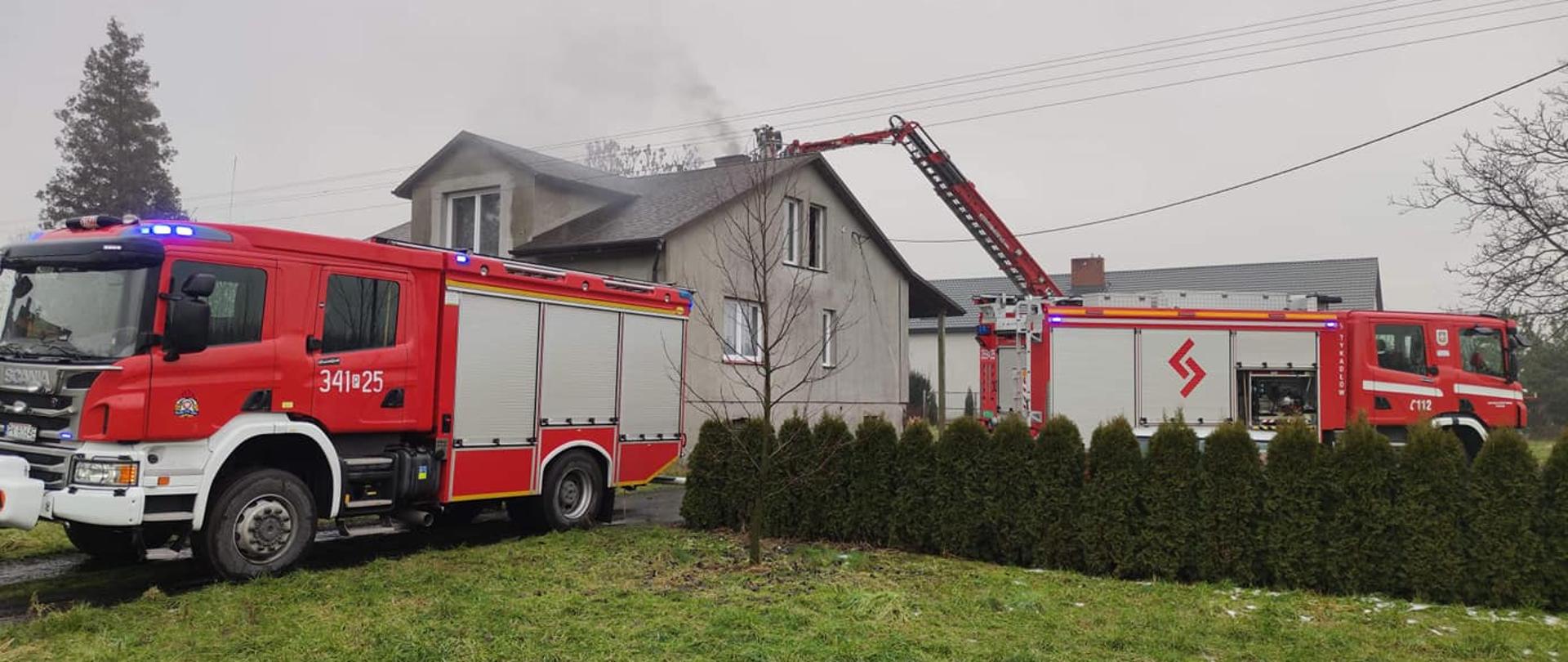 Na zdjęciu widzimy dwa samochody strażackie podczas działań gaśniczych. Z lewej strony kadru samochód strażacki marki Scania o numerze operacyjnym 341-25 skierowany tyłem do budynku, w którym zapaliły się sadze w kominie. Z prawej strony kadru widzimy drugi samochód strażacki również zwrócony tyłem do budynku z bocznym napisem TYKADŁÓW. W dalszej części kadru widzimy budynek jednorodzinny w którym zapaliły się sadze, koloru szarego, dwuspadowy dach koloru brązowego. Nad dachem budynku widzimy rozłożony podnośnik strażacki a w nim strażaków, którzy gaszą sadze w kominie. Przed pojazdami widzimy ogród, zielona trawa, w rzędzie nasadzone niewielkie tuje. 