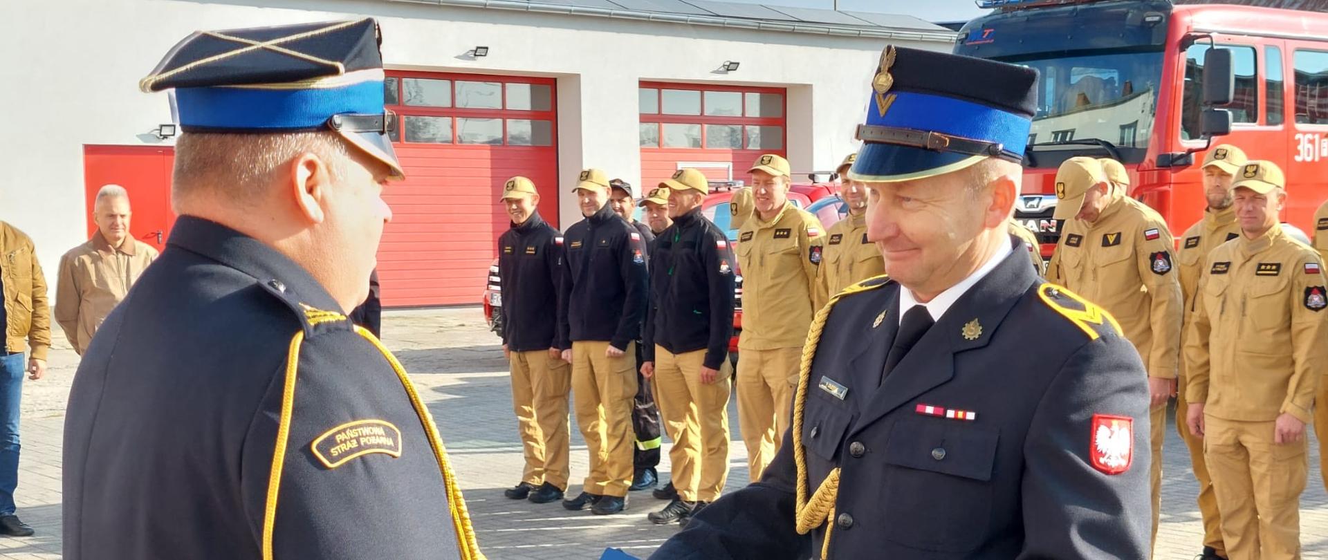 Na zdjęciu strażak Państwowej Straży Pożarnej w umundurowaniu galowym odbiera decyzję o zaopatrzeniu emerytalnym. Uroczysty moment podsumowujący wieloletnią służbę, pełną poświęcenia i oddania dla bezpieczeństwa mieszkańców.