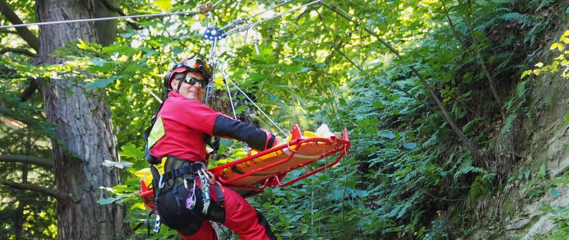 Na zdjeciu widoczny jest ratownik wysokościowy w czerwonym kombinezonie wyposażony w sprzęt ratownictwa wysokościowego. Ewakuuje poszkodowanego na noszach. W tle tereny leśne.