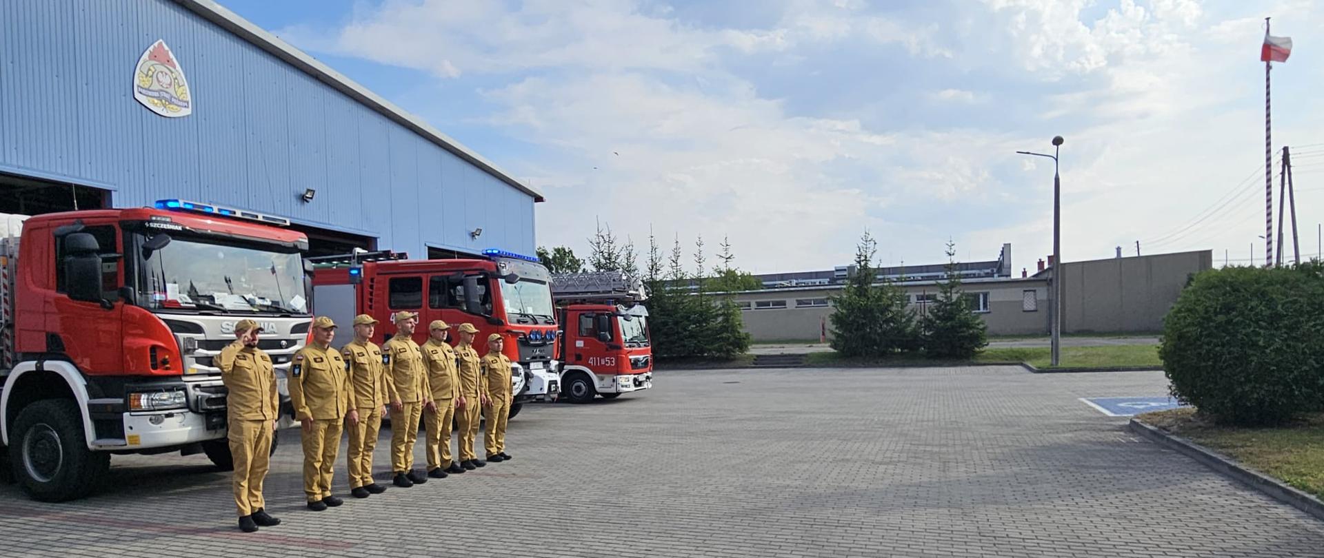 Zdjęcie obrazuje strażaków w umundurowaniu koszarowym, którzy przed garażem jednostki ratowniczo-gaśniczej oddają hołd bohaterom Powstania Warszawskiego. W otwartych bramach garażowych widać samochody ratowniczo-gaśnicze z włączonymi sygnałami świetlnymi. Jeden ze strażaków oddaje honor.
