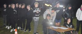 Biwak klas mundurowych. Zdjęcie przedstawia teren biwaku, na którym odbywa się nocny sprawnościowy tor przeszkód. Uczestnicy w kolejce oczekujjąoją kolejność startu. Przy stoliku siedzi policjant. Z prawej strony widoczny jest fragment pojazdu pożarniczego.
