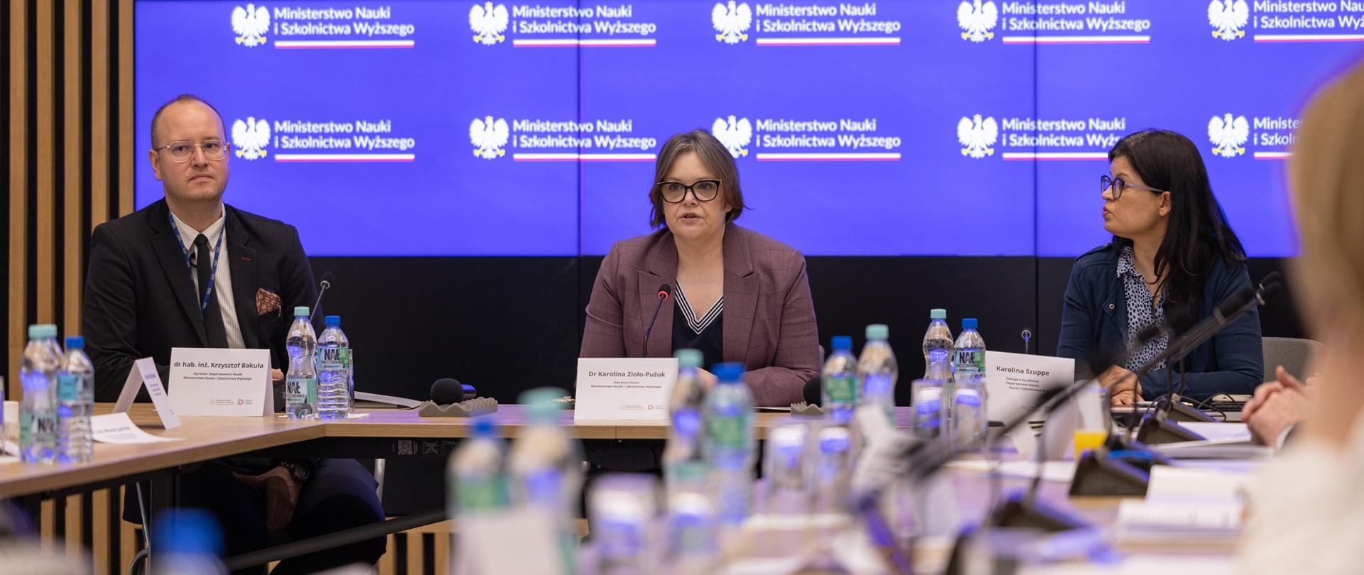 Pod wbudowanym w ścianę z cienkich pionowych listewek wielkim ekranem z napisem Ministerstwo Nauki i Szkolnictwa Wyższego za drewnianym stolikiem siedzi pomiędzy mężczyzną w garniturze i kobietą w granatowym sweterku wiceminister Zioło-Pużuk i mówi do małego mikrofonu.