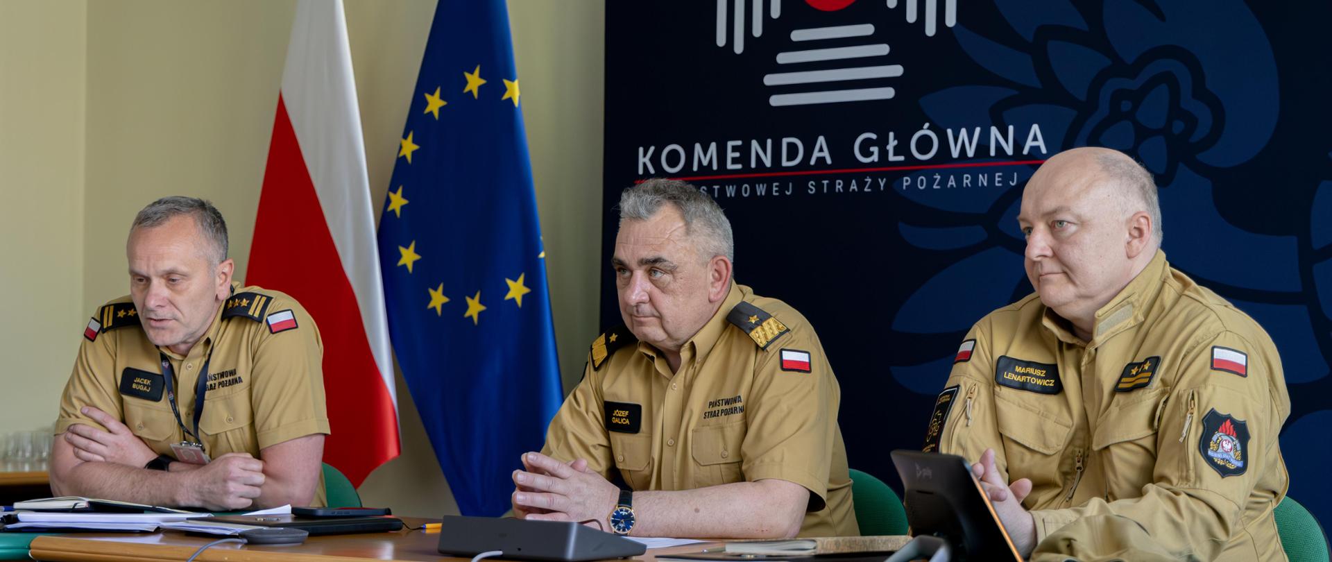 Trzech funkcjonariuszy PSP siedzi przy stole, w tle flaga polski i unii europejskiej.