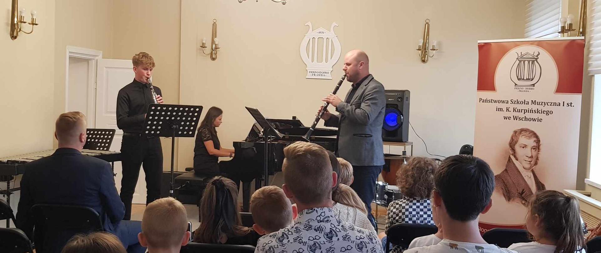 Występ duetu klarnetowego z akompaniamentem w auli szkoły. Widoczna część publiczności.