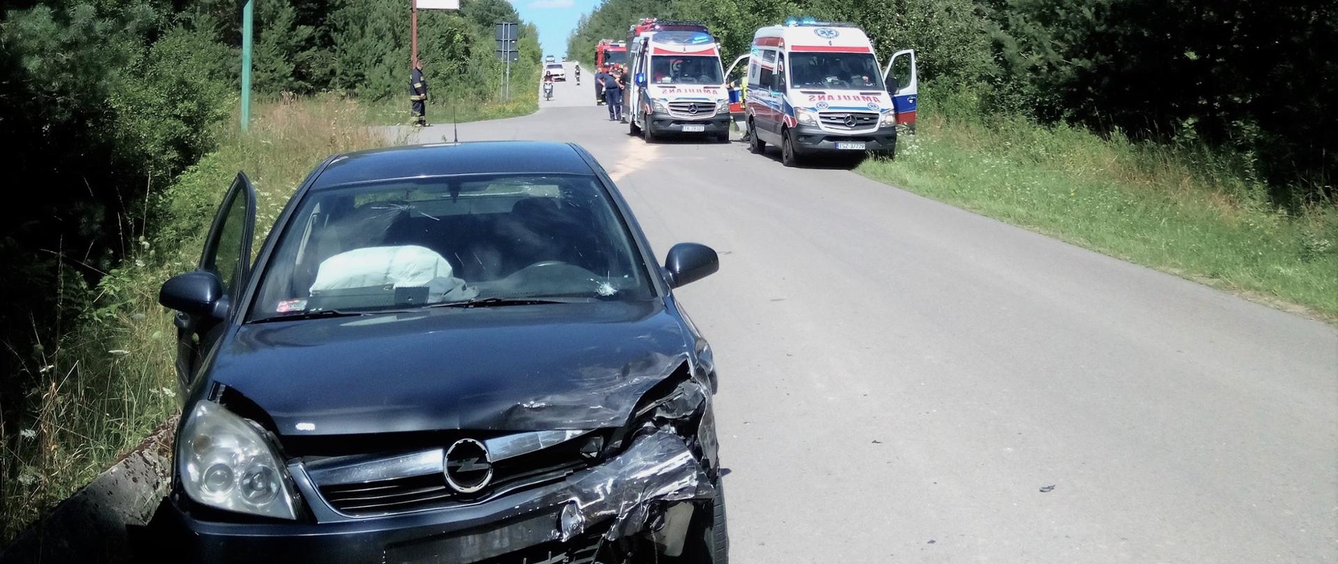 Zdjęcie przedstawia samochód osobowy, który uległy zderzeniu. W oddaleniu, na ulicy widać zaparkowane dwa ambulanse Państwowego Ratownictwa Medycznego.