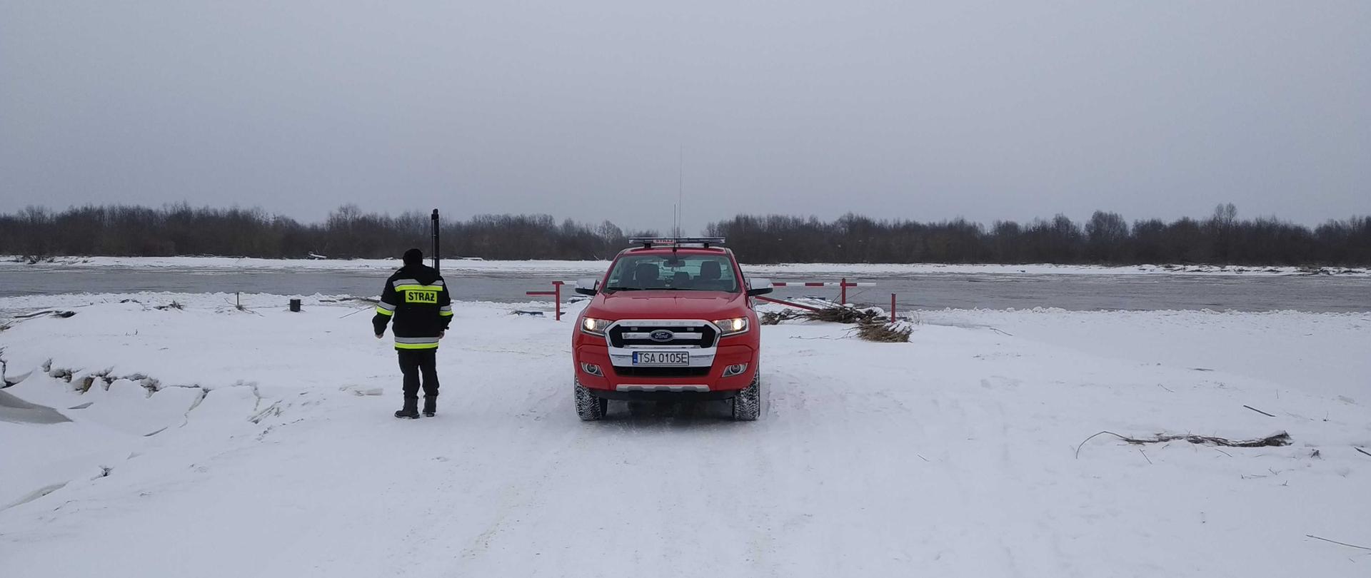 Zdjęcie zrobione na ośnieżonym brzegu rzeki Wisły w miejscowości Zawichost na przeprawie promowej. Na zdjęciu widać samochód strażacki i obok niego strażaka, który wykonuje rekonesans stanu wałów przeciwpowodziowych i zalodzenia rzeki Wisły 