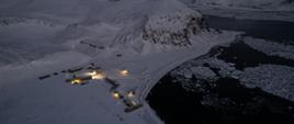 Widok z lotu ptaka na oświetlony kompleks budynków stacji, wszędzie śnieg, z boku widać czarną zatokę pokrytą lodową krą.