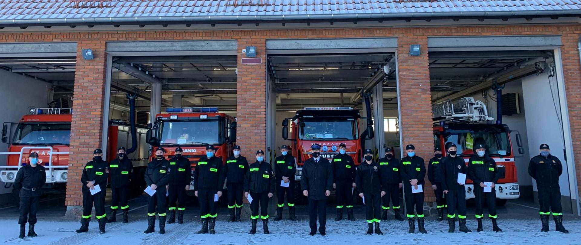 Zdjęcie przedstawia absolwentów szkolenia dowódców OSP oraz członków komisji egzaminacyjnej na tle pojazdów pożarniczych w garażach obiektu KP PSP w Gołdapi
