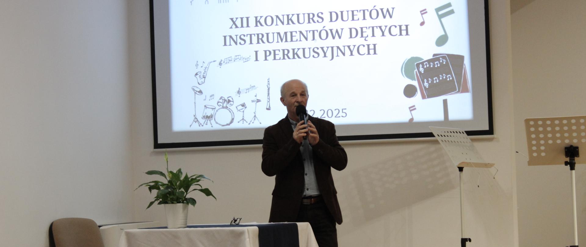 organizator konkursu duetów przemawia do mikrofonu, stoi przy projektorze z wyświetlanym plakatem, w tle głowy publiczności