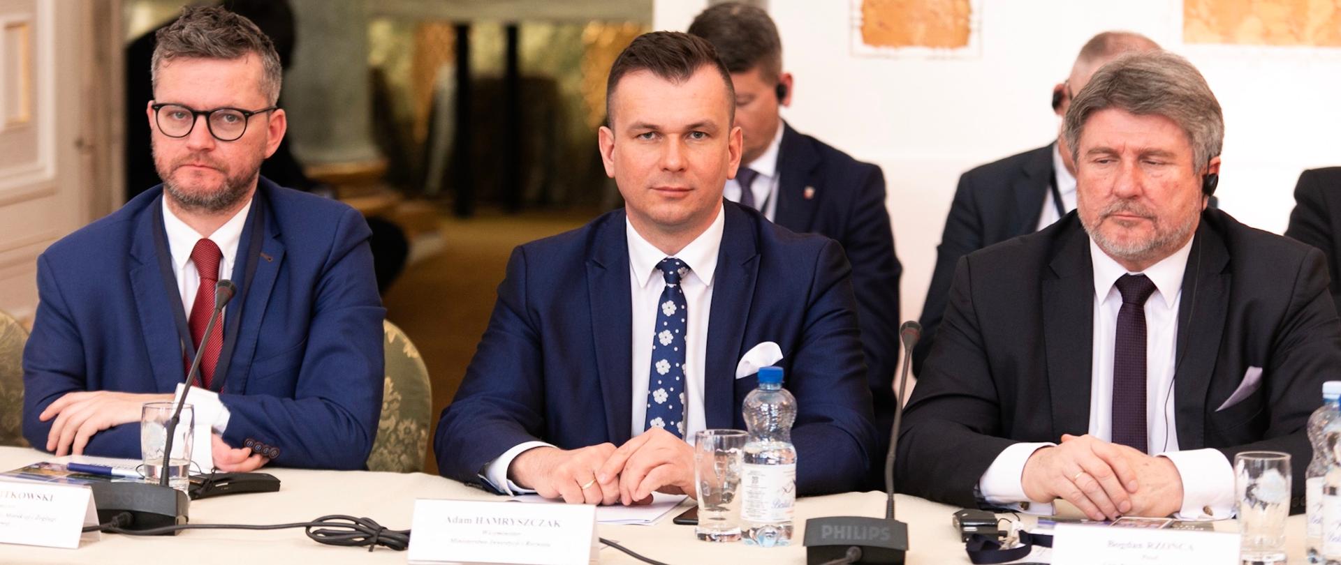 Na sali przy stole siedzą uczestnicy konferencji. Po środku siedzi wiceminister Adam Hamryszczak.