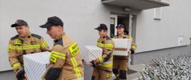 Zdjęcie wykonano w ciągu dnia przed wejściem do budynku Komendy Powiatowej Państwowej Straży Pożarnej w Sępólnie Krajeńskim. Czterech strażaków Państwowej Straży Pożarnej ubraniach specjalnych niesie paczki świąteczne.